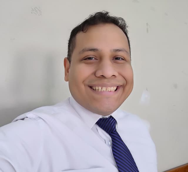 Homem com cabelo curto e escuro, com um largo sorriso. Usa uma camisa branca e uma gravata azul, posando de forma formal.